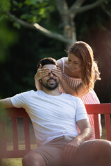 Couple in love, woman covers her boyfriend's eyes from behind on a bench