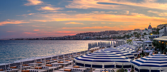 Blue and white striped umbrellas on a pebbled beach, panorama of a golden sunset over Mediterranean Sea along the Promenade des Anglais, Nice, France
