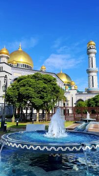 The golden dome and building of the Jamil Asra Hassan Al-Bolkiah Mosque in Bandar Seri Bagawan, Brunei Darussalam, are framed by tropical palm trees on a sunny day. Asia. 4К