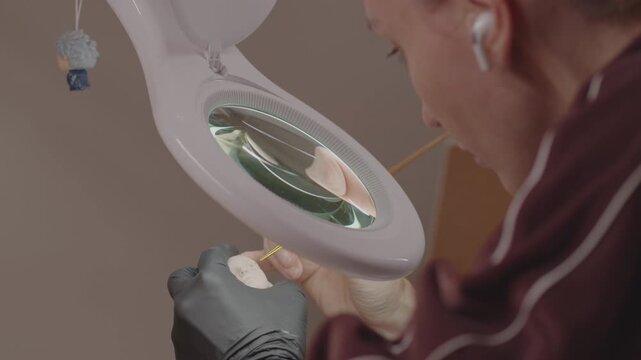 Focused artist using magnifying lamp and fine brush to paint delicate facial details on doll head in workshop