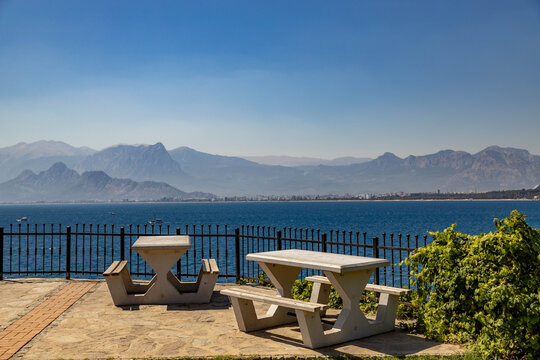 Scenic landscape of the Mediterranean sea and Taurus mountains in the haze. Concrete picnic tables on a stone terrace overlooking the bay. Sunny morning, September, Antalya, Turkey.