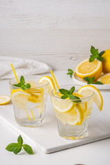 Fototapeta premium Refreshing drinks with lemon slices and mint leaves on a white surface in a bright setting and fresh fruits in the background