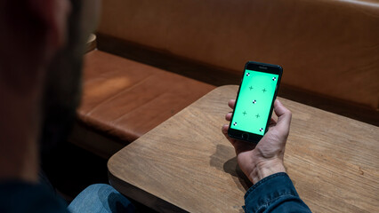 A mature man sitting at a cafe, holding and looking at a smartphone with a green screen chroma key. Perfect for app mockups, website previews, or digital content replacement