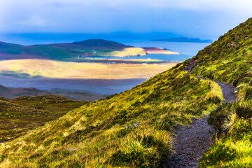 Dramatic hillside trail overlooking coastline on Isle of Skye, green highlands descending toward the sea, atmospheric Scottish landscape with depth and natural light, travel and hiking concept.