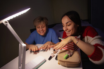 Smiling mother and adult daughter applying nail polish together during home manicure under desk lamp. Cozy evening self care, family bonding and beauty routine at home.