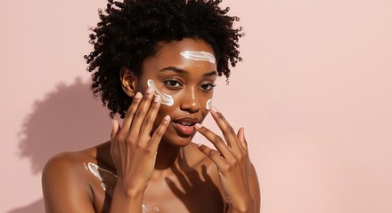 Young black woman applying face cream during her skincare routine. Portrait of african american model with glowing skin using cosmetic product. Self-care and beauty concept on pink background