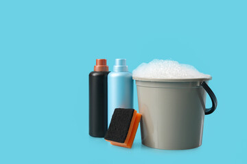 Plastic bucket with foam, sponge and bottles of detergents on blue background