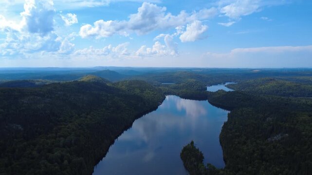 A breathtaking panoramic aerial landscape, revealing the winding mirror lakes of a lush forest with wind turbines in the background under a brilliant blue sky dotted with cottony clouds.