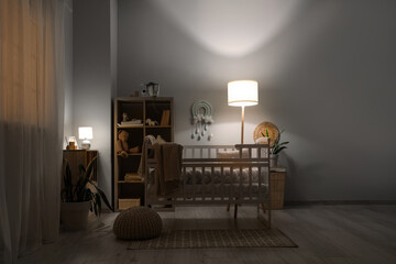 Interior of children's bedroom with baby crib, shelving units and glowing lamps near white wall at...