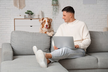 Young man with modern laptop sitting on sofa and cute Australian Shepherd dog in living room