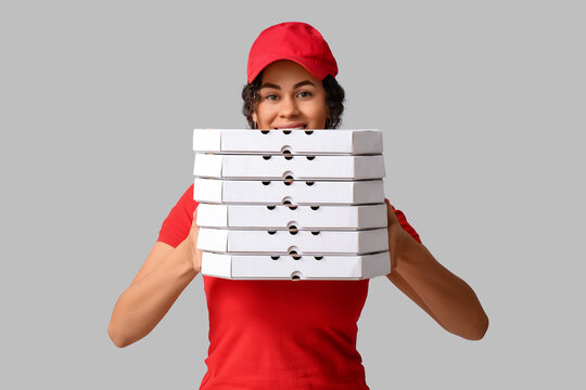 Female African-American courier holding cardboard boxes with tasty pizza on grey background