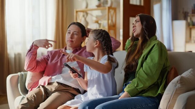 Curious daughter pointing finger watching tv on sofa with mother father closeup