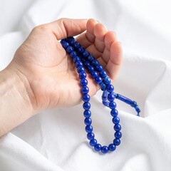 Close Up Of A Hand Holding A String Of Blue Prayer Beads Against A White Fabric Background In Soft Natural Light