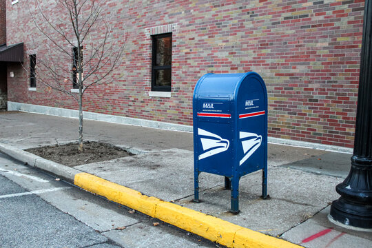 A United States Postal Service (USPS) collection box, commonly known as a "blue box", on a city street with a brick building in the background (Jefferson City, MO - Dec. 25, 2025). Editorial Use Only.