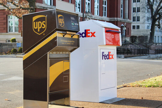 Outdoor UPS and FedEx drop boxes on a city street with a brick building facade in the background and glimpses of bare trees (Jefferson City, MO - Dec. 24, 2025). Editorial Use Only.