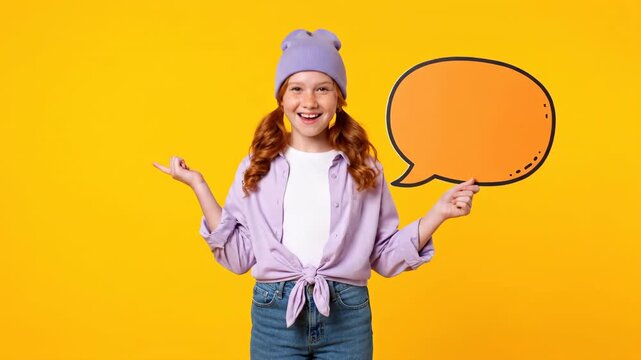 A smiling freckled redhead holds an orange speech bubble on a vibrant yellow background wearing casual denim and lilac. Studio shot emphasizing a cheerful message.