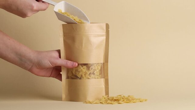 Woman putting raw pasta into stand-up pouch on beige background, closeup
