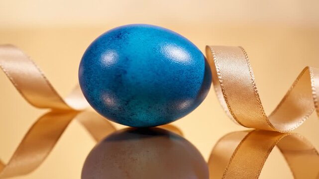 Displaying Single Blue Easter Egg With Gold Ribbon on Reflective Surface