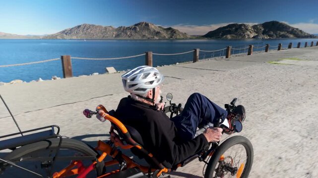 An elderly man enjoys biking on a recumbent electric tricycle by the lake with mountains in the background during a clear day.