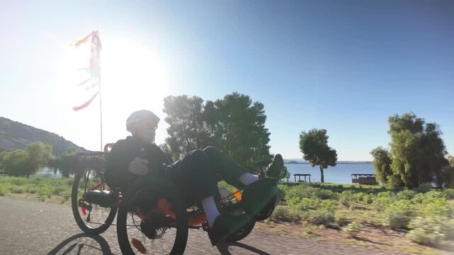 An elderly man rides a recumbent electric trike along a lakeside path under a sunny sky, enjoying his time outdoors.