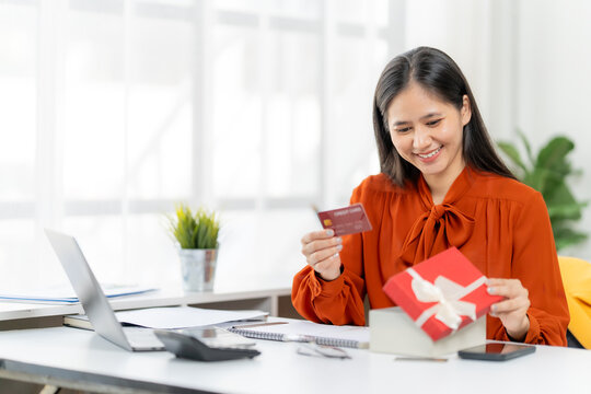 Online Shopping Delight: An Asian woman, radiant with anticipation, holds a credit card while opening a beautifully wrapped gift box, epitomizing the ease and joy of online shopping.