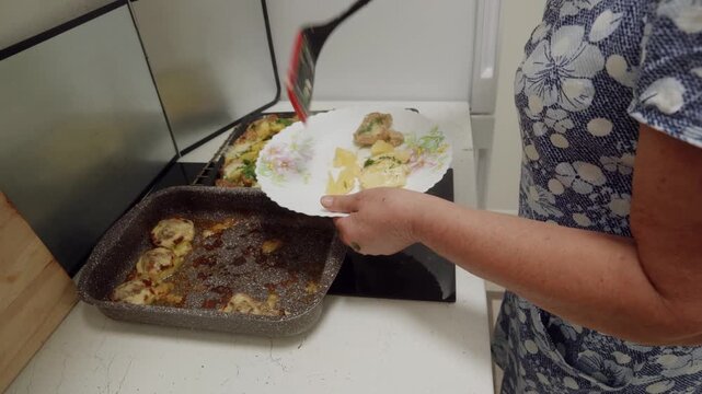 Senior caucasian woman placing portion of baked meat and potatoes onto plate near stove using spatula, concept of home cooking and serving dinner.