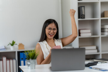 Excited Professional Celebrates Success: A young professional celebrates her success, punching the air in jubilation while working at her desk, as a laptop sits open in front of her.