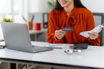Online Transaction: A woman in an orange blouse smiling while holding a credit card and receipts, surrounded by a laptop and calculator, ready to begin the payment process.
