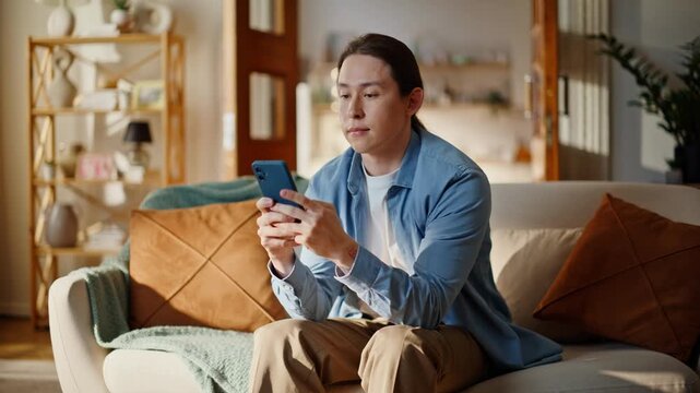 Asian guy typing smartphone looking screen at couch comfortable house closeup