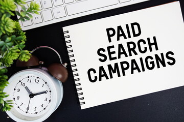 Paid Search Campaigns text written on notepad with keyboard and alarm clock on black background