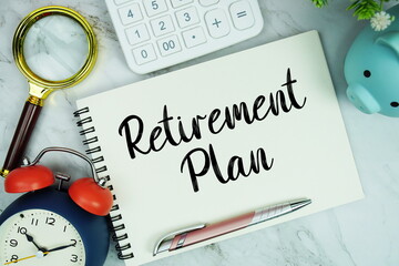 Retirement Plan text written on notepad with piggy bank, calculator, magnifying glass and alarm clock on marble background