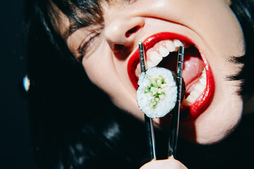 Bold woman with red lips bites sushi roll, expressing playful enthusiasm and passion for food, on a dark background emphasizing vibrant colors.