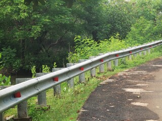Guardrail with red reflectors on a rural roadside with lush greenery and a dirt shoulder in a natural setting.