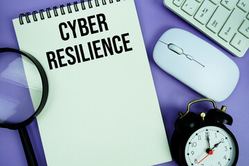 Notepad with the text "Cyber Resilience" and white keyboard, wireless mouse, magnifying glass and alarm clock