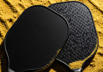 Two black pickleball paddles with textured surfaces on vibrant yellow sand