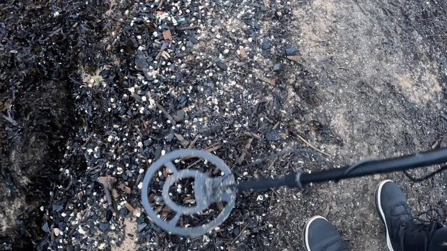 A metal detector coil sweeps across a dark, shell-covered shoreline as the searcher scans through washed-up debris and mixed textures.