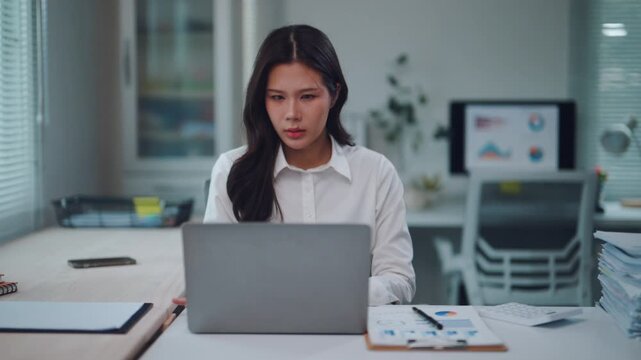 Young Asian businesswoman feeling stressed and tired while working late at night in the office. Busy woman managing paperwork and data analysis