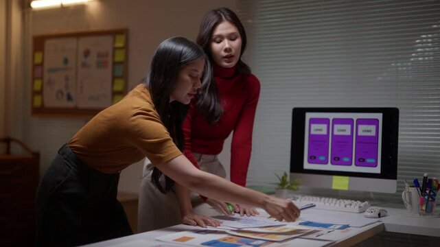 Two young Asian women collaborating on a UX project. Creative workers discussing mobile application wireframes and planning strategy in a dark office