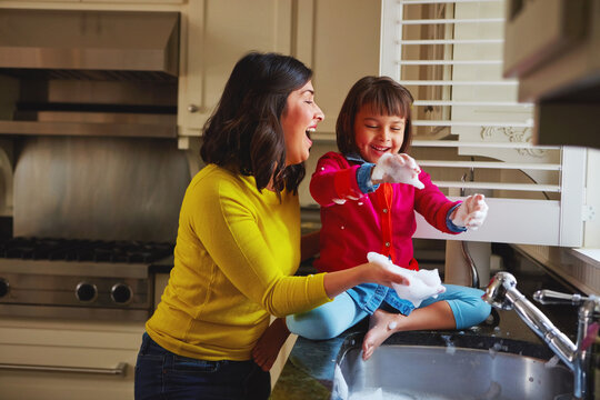Laugh, mother and child with foam at kitchen sink for washing dishes, love or healthy relationship. Funny family, mom or kid playing with dish soap for cleaning support, fun chores or bonding in home
