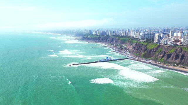 Sunny morning Miraflores Barranco Barranquito Playa Makaha beach pier aerial drone Lima Peru clear hazy blue skies residential skyscraper buildings cars traffic surf waves ocean seaside forward motion