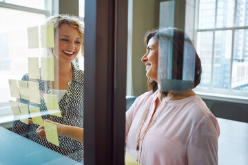 Glass wall, women and happy with sticky notes in meeting for story approval, guidance or workflow. Editor, journalist or discussion in newsroom for article advice, news development or task management