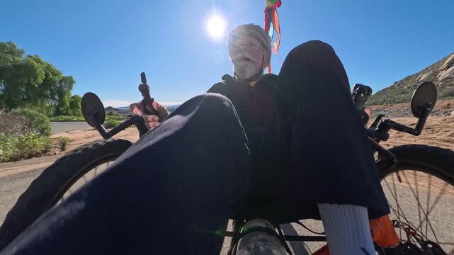 Elderly man enjoys biking on a recumbent electric tricycle under bright sunlight in a natural setting.