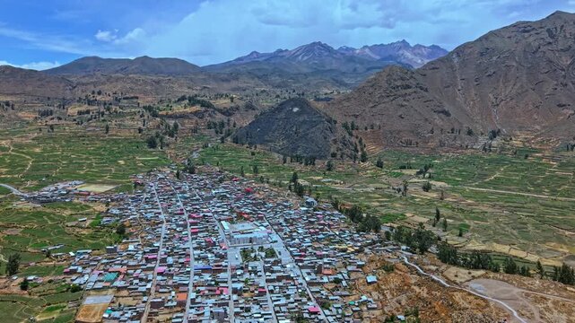 Aerial drone footage flies toward and over the town of Cabanaconde, highlighting terraced farmland, colourful Andean mountains, and the village&rsquo;s rural setting in the Colca Valley, Peru.
