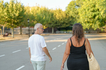 Fototapeta premium A man and a woman are walking down a street. The man is wearing a white shirt and the woman is wearing a black dress. They are both carrying handbags. The scene is peaceful and calm