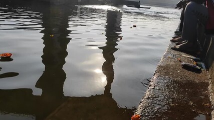 Static shot of devotees offering floating lamps during deepdaan ritual, reflections of pillars and sunlight shimmering on calm river water.