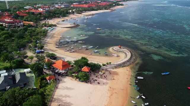 Aerial drone footage of shallow coastal water along a curved sandy beach with small boats near shore, clear reef patches a stone breakwater and coastal infrastructure forming a busy tropical shoreline