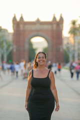 A woman in a black dress is smiling in front of a large archway. The scene is lively and bustling with people walking around