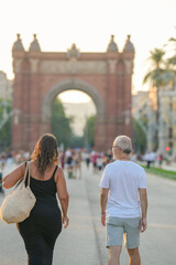 A woman and a man walk down a street with a large arch in the background. The woman is carrying a handbag and the man is wearing a white shirt and shorts. The scene is lively
