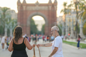 A man and a woman are walking down a street in front of a large arch. The man is holding a woman's hand and they are both smiling. The scene is lively and cheerful, with many people walking around