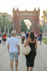 A man and a woman walk down a street in front of a large arch. The man is wearing a white shirt and shorts, while the woman is wearing a black dress and a straw bag. The scene is lively and bustling